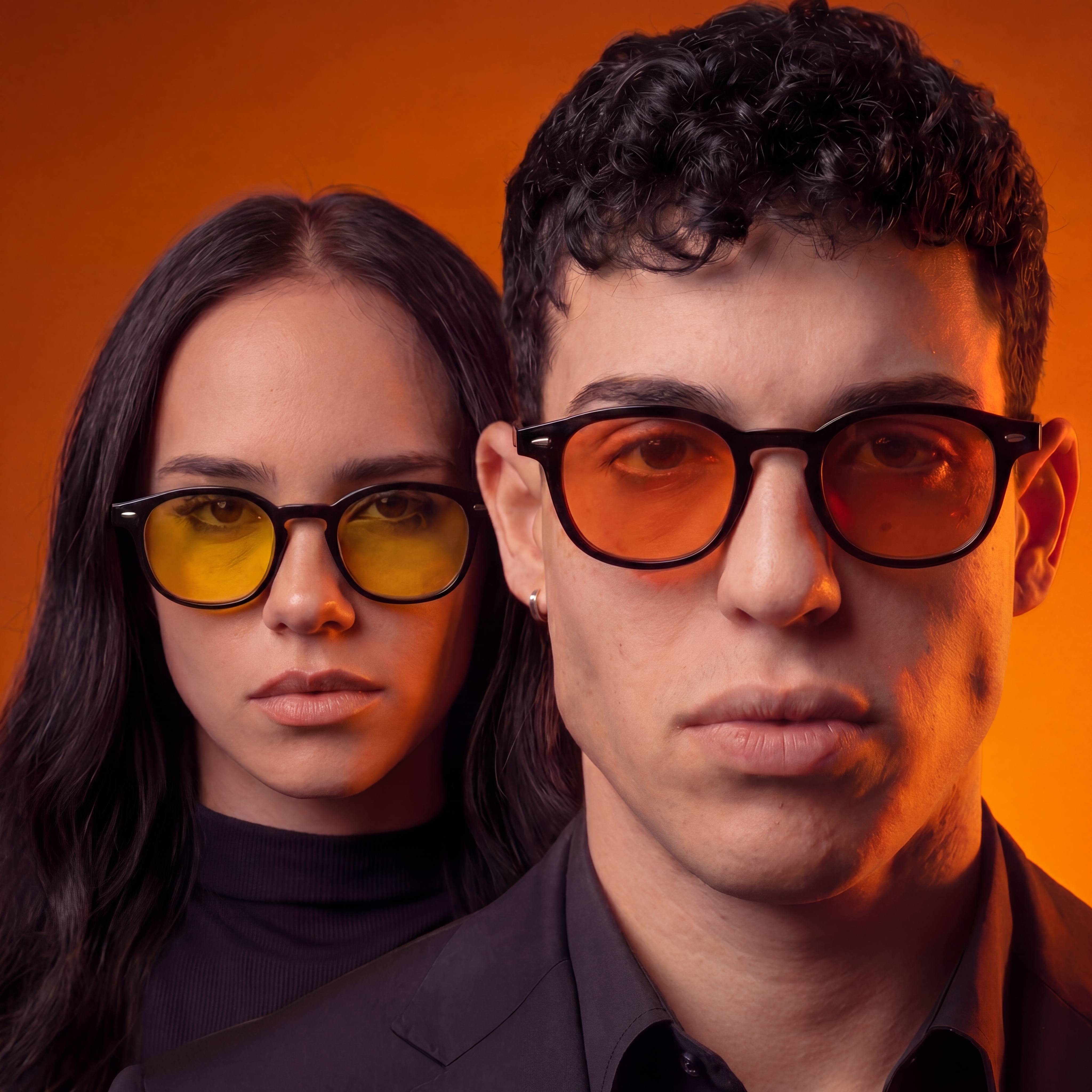 A studio portrait of a young man with dark curly hair wearing yellow-tinted glasses and a black suit.